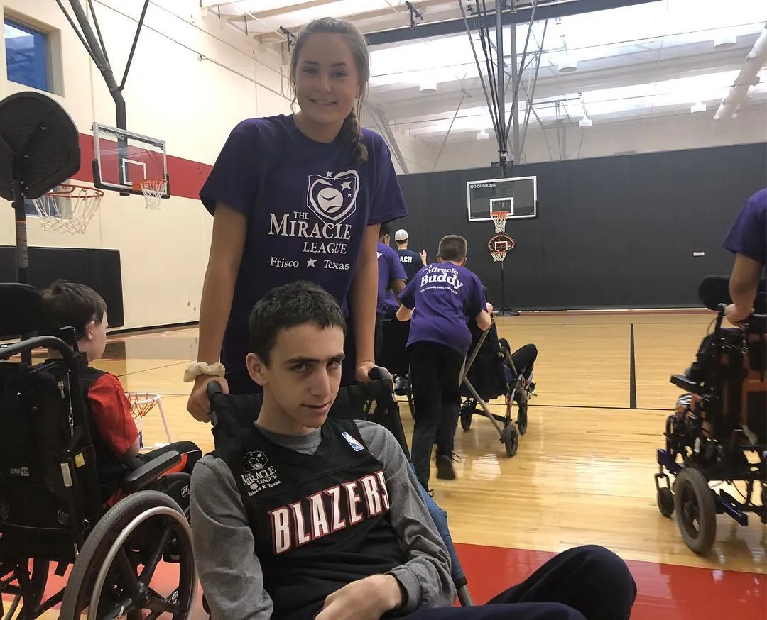A volunteer in a purple Miracle League shirt stands behind a boy in a wheelchair on a gym floor