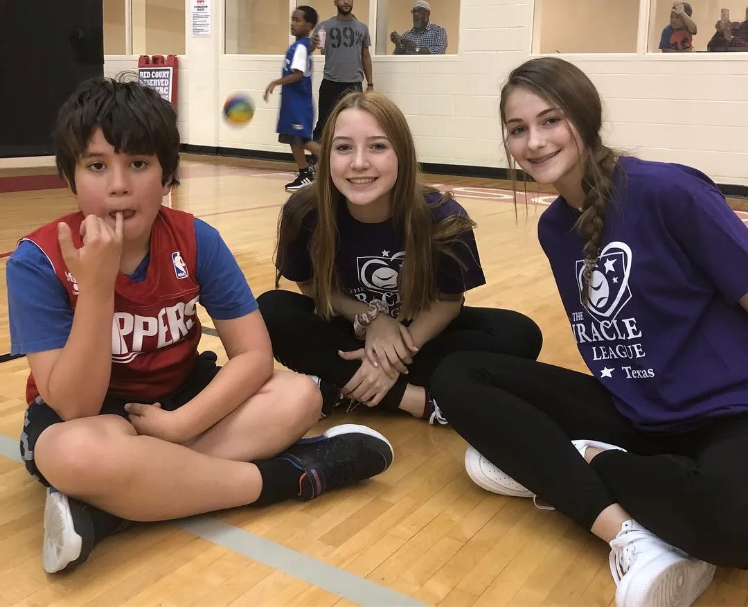 A boy in a red jersey sits on a gym floor beside two smiling girls in purple Miracle League shirts