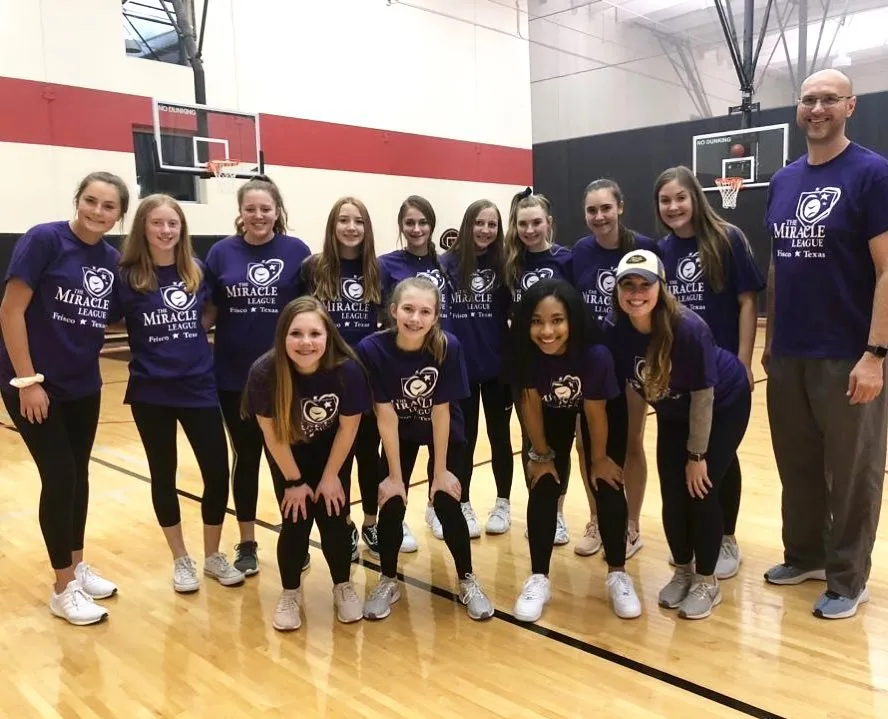 A group of girls and a man in purple Miracle League Frisco Texas shirts pose together in a gym