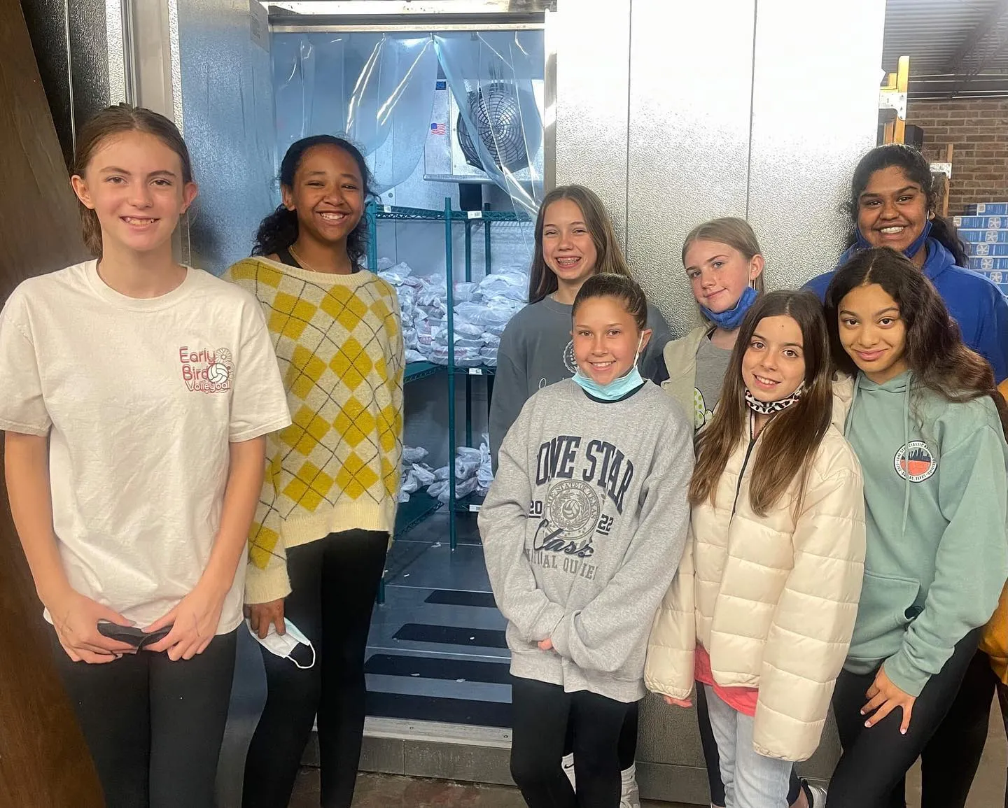 Eight smiling girls stand in front of an open walk-in cooler