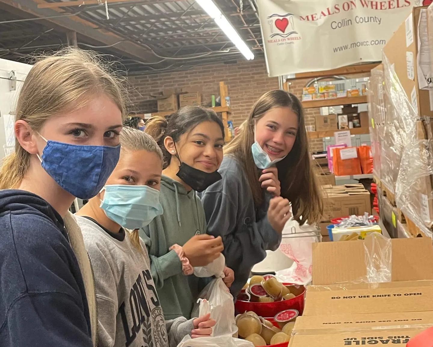 Four girls in face masks packing food at a Meals on Wheels Collin County warehouse