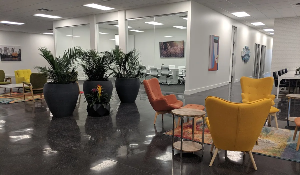 Lounge seating in orange and yellow wing chairs beside tall plants, with glass meeting rooms behind